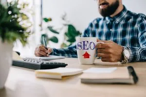 cropped shot of successful smiling seo developer with cup of coffee and computer writing notes at
